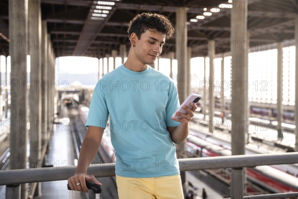 A young man in a blue t-shirt stands at a train station, holding a suitcase and using his smartphone. Bright sunlight illuminates the modern platform and tracks