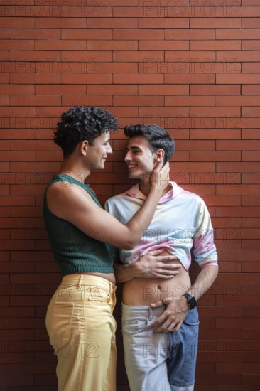 A joyful gay couple embracing and smiling at each other, standing against a red brick wall. The tender moment captures love and connection in an urban setting