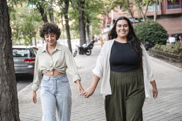 A lesbian couple is walking hand in hand on a city street, smiling and enjoying a sunny day. The scene captures warmth, happiness, and togetherness