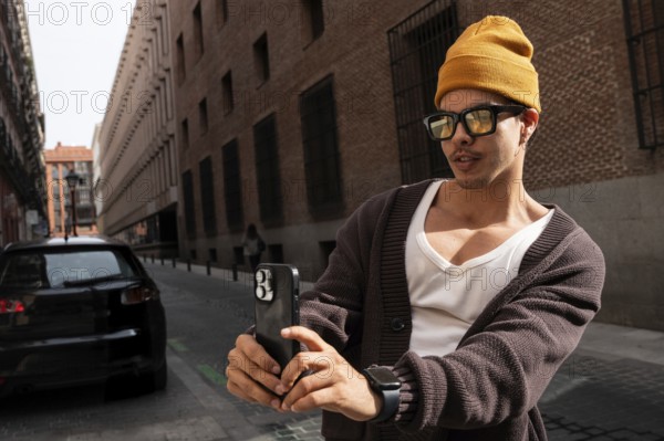 A stylish latin man wearing glasses and a yellow beanie takes a picture with his smartphone on a sunlit city street, showcasing urban fashion and modern technology
