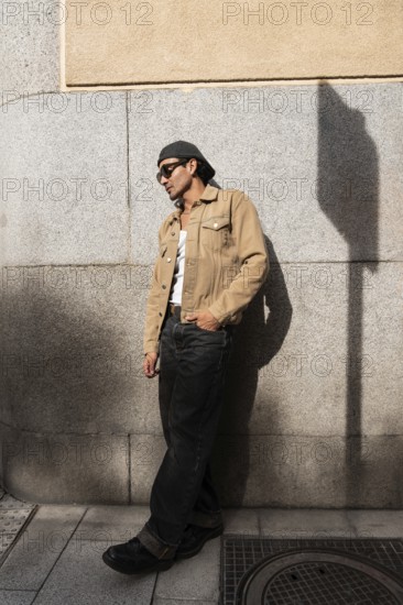 A man stands casually in front of a stone wall, wearing sunglasses, a flat cap, a beige jacket, and a white tank top. His relaxed pose and attire give off an urban vibe