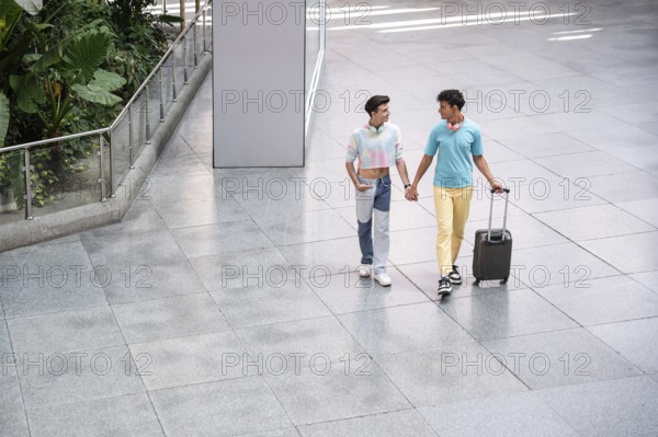 A joyful gay couple walks hand in hand through an airport terminal with luggage, enjoying each other's company and looking stylish in casual clothing