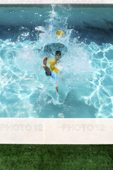 A young boy in a yellow shirt and blue shorts jumps energetically into a bright blue swimming pool, creating a large splash. The scene captures a moment of fun and summer joy