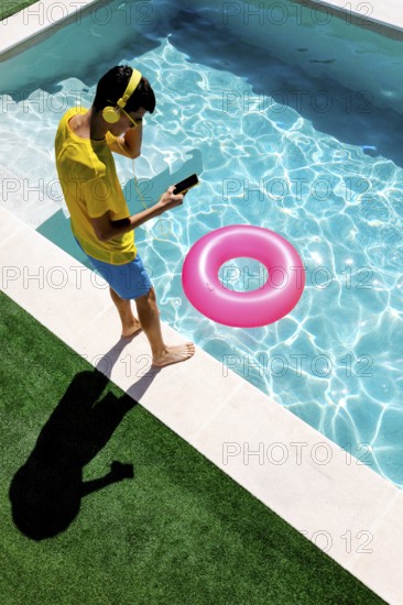 A person in vibrant attire enjoys music with headphones at a pool. Next to them, a pool float adds a splash of color to the clear blue water, capturing a moment of summer relaxation