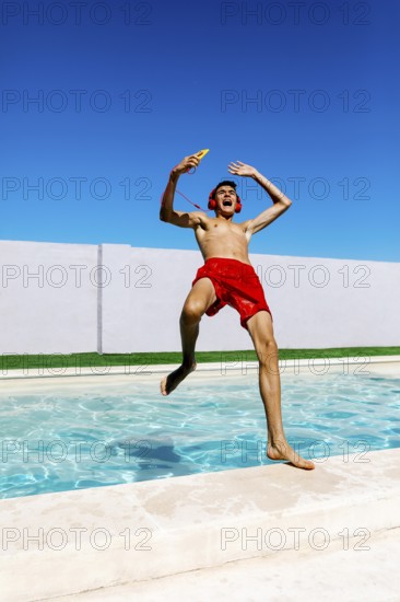 A joyful young teenage boy in red trunks leaps into a pool, wearing headphones and holding a popsicle, against a vivid blue sky, capturing the essence of carefree summer fun