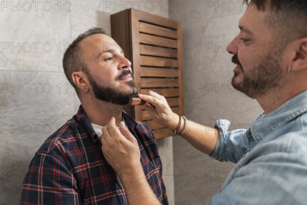 A gay couple shares a tender grooming moment in their bathroom. One helps the other style his beard with care, showcasing their connection and trust