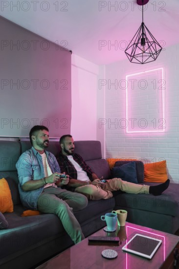 A gay couple relaxes on a sofa, playing video games in a warmly lit living space with modern decor. The room features a stylish lamp and neon accents