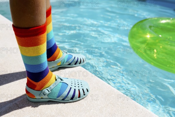 A vibrant scene featuring rainbow-striped socks paired with blue sandals by a bright swimming pool. The colorful socks and green inflatable ring add a playful summer vibe