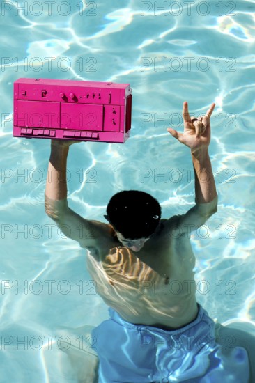 A person in blue shorts enjoys a sunny day in a pool, holding a vibrant pink boombox above the water. The scene captures a lively and fun summer atmosphere