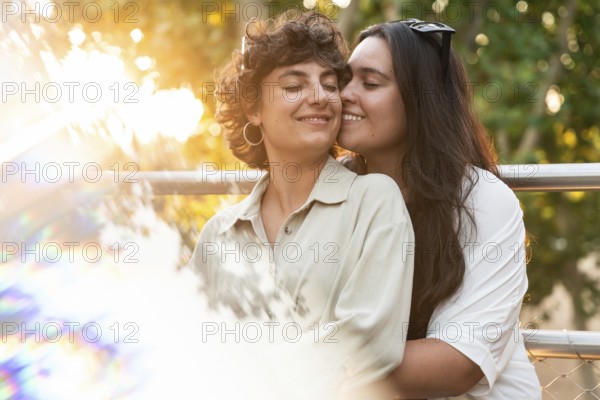 A loving lesbian couple shares a tender embrace outdoors, surrounded by vibrant nature. The sunlight adds a warm and serene ambiance to their affectionate moment