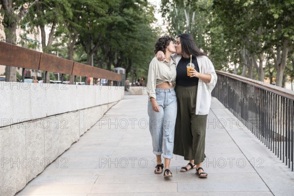 A lesbian couple shares a tender kiss while walking down a city street, displaying affection and happiness. They enjoy a refreshing drink amidst the greenery and urban setting