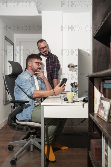 A gay couple collaborates at home, with one seated and the other standing, reviewing a smartphone. The bright room creates a cozy, productive atmosphere