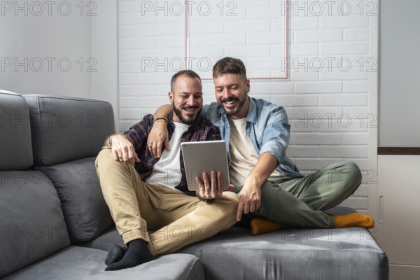 A joyful gay couple is comfortably seated on a sofa, engaged with a tablet. Their smiles reflect happiness and a strong connection, creating a warm and inviting atmosphere at home