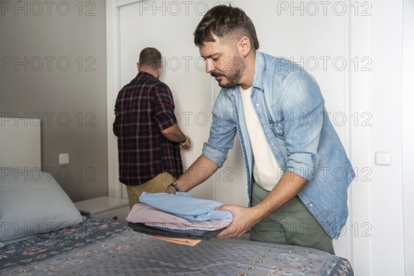 A gay couple organizes clothes and tidies up their bedroom. One is folding laundry on the bed while the other is walking towards a closet. The room is bright and inviting