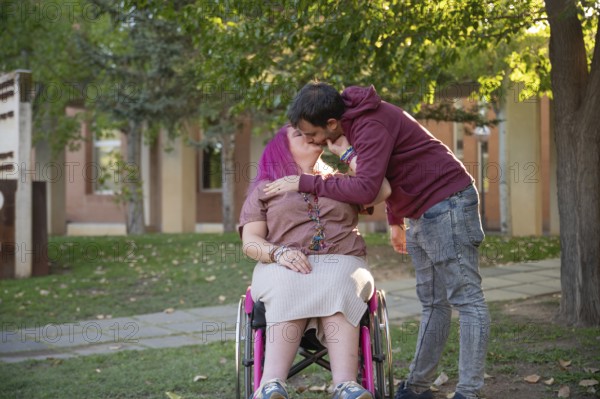 A couple shares an affectionate kiss on a peaceful college campus The woman, with pink hair and spina bifida, sits in a wheelchair, embraced by her partner under the trees