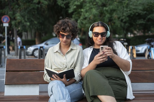 A lesbian couple enjoys a sunny day outdoors, sitting on a bench. One is reading a book, and the other listens to music on headphones, both wearing sunglasses