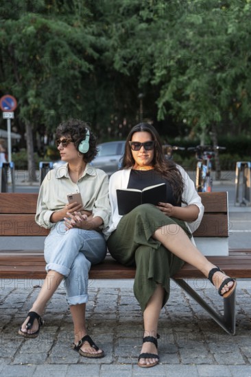 Lesbian couple relaxes on a park bench. One is reading a book, while the other listens to music, phone in hand. Surrounded by greenery and enjoying the tranquil setting