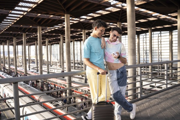 A joyful gay couple at a modern train station, sharing a moment with a smartphone, ready for their journey. Stylish attire and a luggage set the scene for an adventure