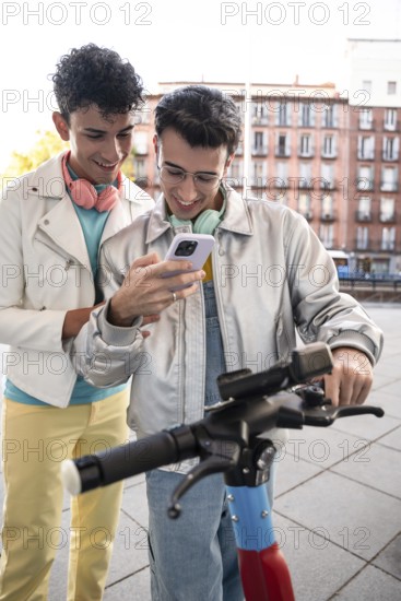 A happy gay couple, each wearing headphones, enjoys a light-hearted moment outdoors while looking at a smartphone to rent a electric scooter