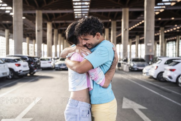 A joyful gay couple shares an embrace in a spacious urban parking garage, surrounded by parked cars. The warm moment captures love and happiness in a contemporary setting