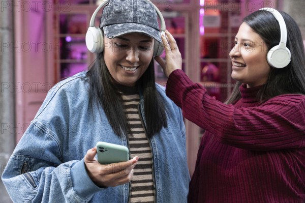 A smiling Latin lesbian couple enjoying music together with headphones. One holds a smartphone, sharing a joyful moment in a vibrant urban setting