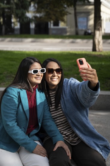 A smiling Latin lesbian couple enjoys a sunny day at the park, embracing while taking a selfie with their smartphone, capturing a joyful moment together