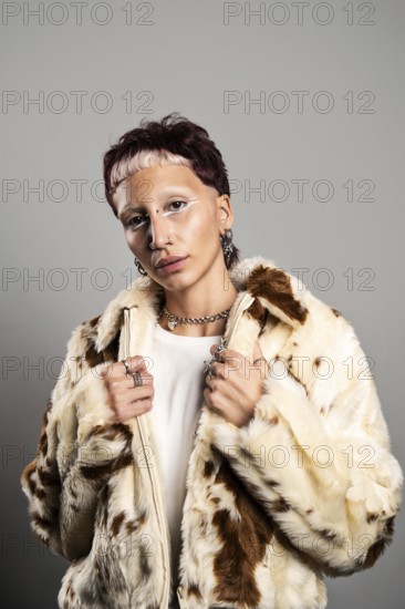 A woman with distinctive makeup and jewelry poses confidently looking at camera in a stylish faux fur coat. The combination of accessories and makeup creates an edgy, modern look