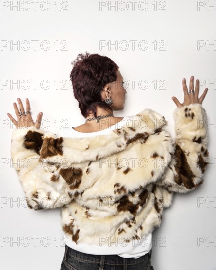 A woman with a trendy mullet hairstyle, adorned with earrings and rings, wearing a stylish cream and brown faux fur jacket against a plain background