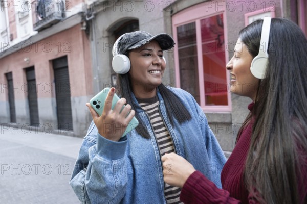 A joyful Latin lesbian couple wearing headphones and smiling while listening to music. They are standing on an urban street, enjoying their time together and sharing music