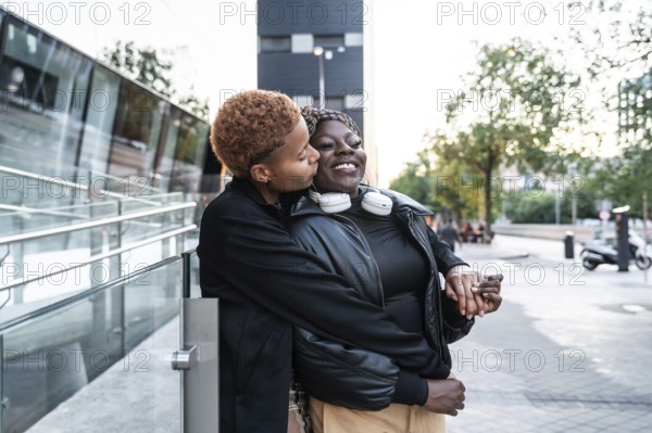 A loving LGBT multiethnic couple is embracing and smiling on an urban street. One partner gently kisses the other's cheek, radiating joy and warmth, against a modern city backdrop