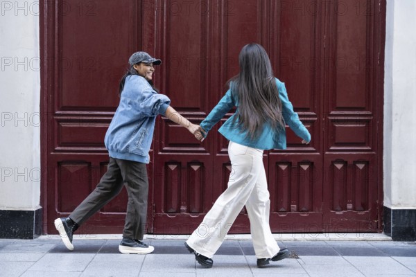A joyful Latin lesbian couple holding hands as they walk together. They are both wearing casual attire and walking in front of a red wooden door, enjoying each other's company