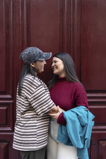A joyful Latin lesbian couple in a loving embrace stands against a deep burgundy wooden backdrop, capturing a moment of connection and affection, dressed in casual attire