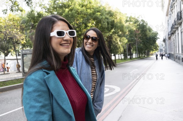 A joyful Latin lesbian couple strolls hand in hand down a tree-lined city street. They wear stylish jackets and sunglasses, embracing their love openly under the sun