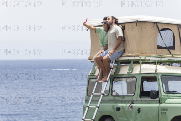 A blind man and a woman sit on the rooftop tent of a motorhome, enjoying a coastal view while taking a selfie. The scene reflects adventure, connection, and outdoor exploration