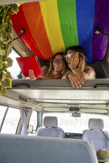 A joyful lesbian couple relaxes in a van under a rainbow flag, sharing laughs and browsing on a tablet. This image embraces love, freedom, and digital connectivity on the road