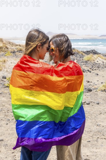 Two women wrapped in a rainbow flag share a moment on a rocky beach, symbolizing love and pride in a picturesque coastal setting with waves in the background