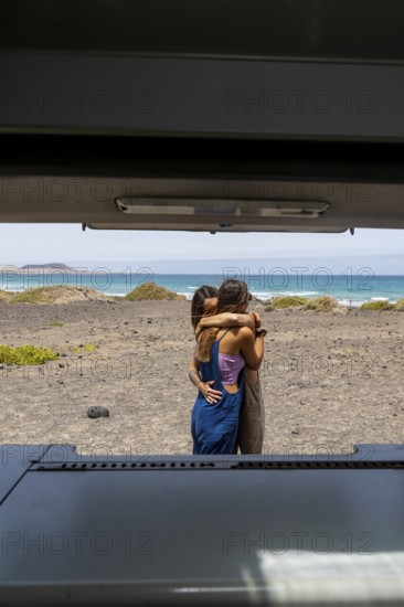 A lesbian couple shares an intimate embrace by the ocean, viewed from inside a camper. The serene beach landscape adds to the feeling of tranquility and adventure