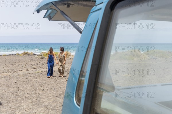 A lesbian couple strolls hand in hand along a beach, adjacent to a classic van. They enjoy the tranquil sea breeze and the picturesque ocean view, creating a scene of adventure and relaxation