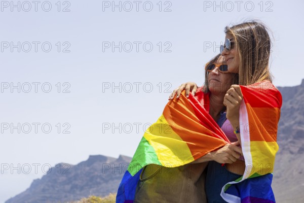 Two women embrace under a colorful rainbow flag, symbolizing LGBTQIA pride and unity, set against a serene outdoor backdrop, representing love and acceptance