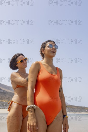 Lesbian women in stylish orange swimsuits enjoying a sunny beach day. They wear sunglasses, showing joy and relaxation with the sea in the background under a clear sky