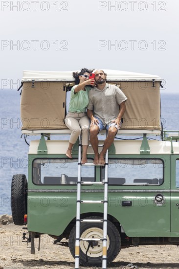 A blind man and a woman sit on the rooftop tent of a motorhome, enjoying a coastal view while taking a selfie. The scene reflects adventure, connection, and outdoor exploration