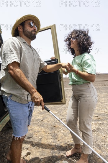A blind man, assisted by a companion, steps out of a motorhome into an arid landscape, experiencing the freedom of travel and adventure. A white cane guides his path