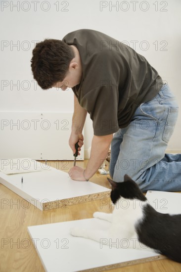 A man assembles new furniture on a hardwood floor with a curious cat watching nearby. He uses tools and follows instructions, focusing on the task at hand