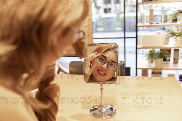 A smiling woman tries on a new pair of eyeglasses while looking at her reflection in a tabletop mirror inside a boutique. The store has a warm and modern ambiance with elegant displays