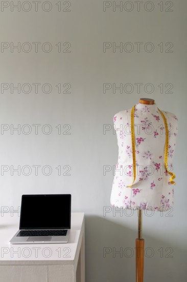 A floral-patterned tailor's mannequin with a yellow measuring tape stands next to a desk with a laptop. The minimalist workspace reflects creativity and fashion design in a modern setting