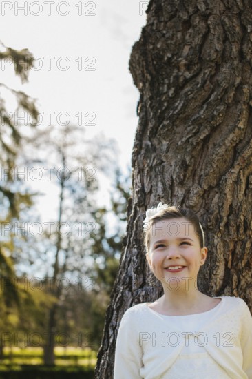 A joyful girl in a white First Communion dress stands beside a tree, beaming with happiness. Sunlight filters through the branches, creating a serene outdoor scene