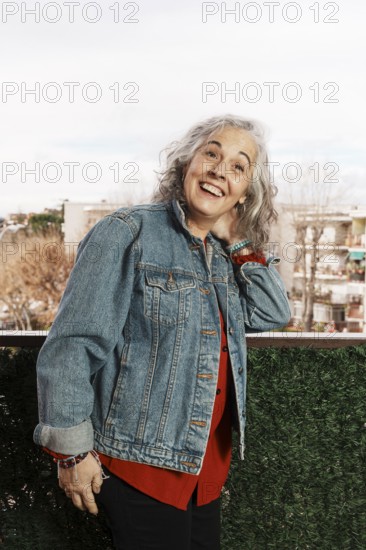 A joyful woman with white hair and a denim jacket smiles brightly while standing on her balcony, surrounded by a pleasant neighborhood view on a clear day