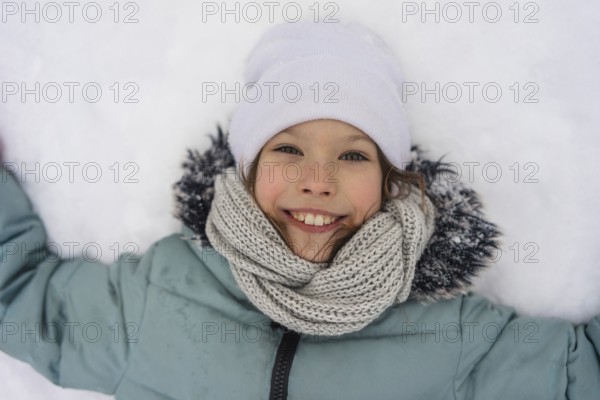 A smiling child lies in the snow, wearing a warm jacket, white hat, and knitted scarf. The joy and warmth of a winter day are captured in this delightful image