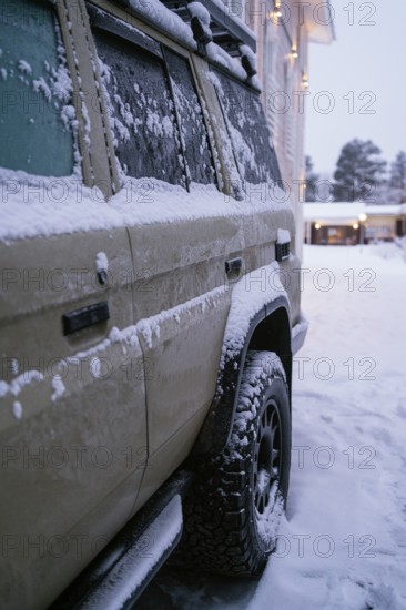A snow-covered vehicle is parked near a warmly lit building in a snowy setting. The scene captures the serene, cold atmosphere of a winter day in Swedish Lapland