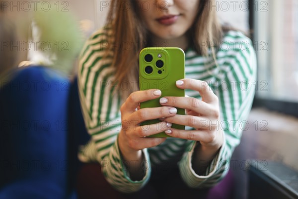 A cropped unrecognizable young woman checks her smartphone while enjoying a coffee break at a cafe. The focus is on a green mobile phone held in her hands